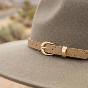 Könnte beinhalten: Nahaufnahme eines grauen Fedora-Hutes mit einem hellbraunen Lederband und einer goldfarbenen Schnalle. Der Hut hat eine breite Krempe und ein klassisches Design, geeignet für verschiedene Anlässe.