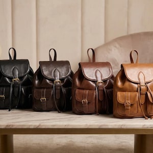 May include: Four leather backpacks in varying shades of brown and black are displayed on a marble-topped table. Each backpack features a flap closure, front pockets, and adjustable straps. The bags have gold-colored buckles and drawstrings.