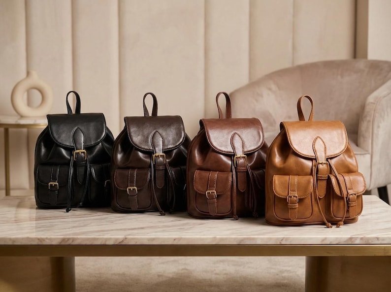 May include: Four leather backpacks in varying shades of brown and black are displayed on a marble-topped table. Each backpack features a flap closure, front pockets, and adjustable straps. The bags have gold-colored buckles and drawstrings.