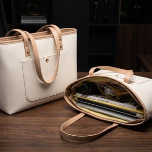May include: Two cream-colored tote bags with tan leather handles and trim sit on a wooden surface. One bag is closed, while the other is open, revealing contents like a laptop and magazines. The bags have a minimalist design.