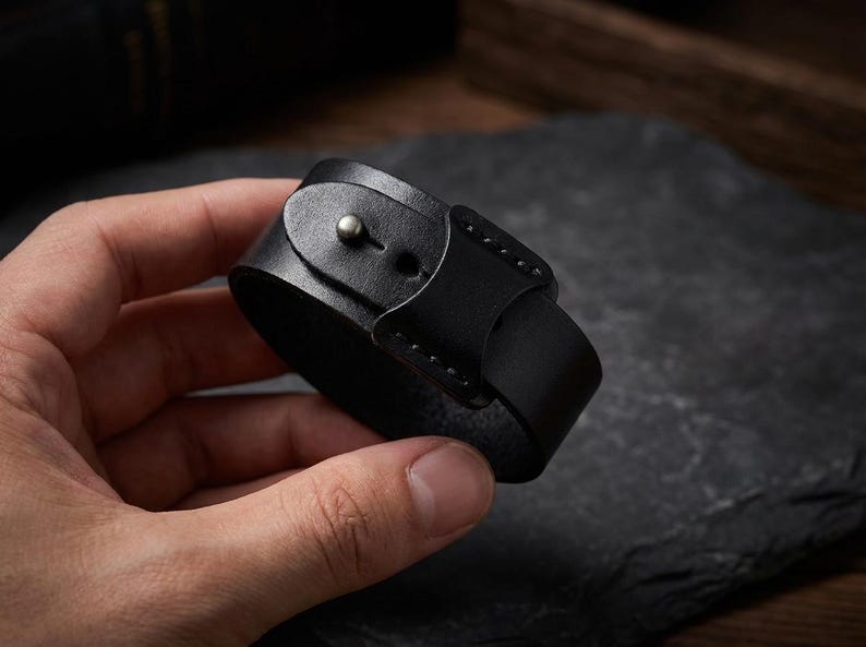 May include: A black leather bracelet with a buckle fastening. The bracelet is made of thick leather and features a silver-coloured metal stud. The design is simple and classic, suitable for everyday wear.