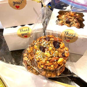 May include: A close-up of a stack of granola bars in a clear cellophane bag. The bars are packed with nuts, seeds, and dried fruit. White boxes with a window and a "Thank you!" sticker are in the background.