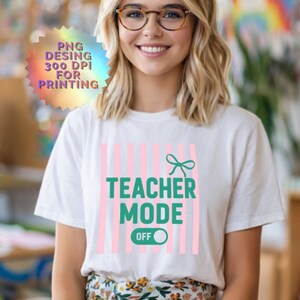 Puede incluir: Una camiseta blanca con un diseño gráfico de rayas verticales rosa claro, un lazo verde azulado y el texto "TEACHER MODE" en letras mayúsculas verde azulado sobre un gráfico de interruptor verde azulado con la palabra "OFF" en blanco.