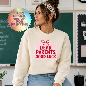 Könnte beinhalten: Ein cremefarbenes Sweatshirt mit Rundhalsausschnitt, das eine rote Schleifengrafik über dem Text "DEAR PARENTS, GOOD LUCK" in roter Schrift vor einem Hintergrund aus vertikalen hellrosa Streifen zeigt. Das Kleidungsstück hat lange Ärmel und einen gerippten Ausschnitt.