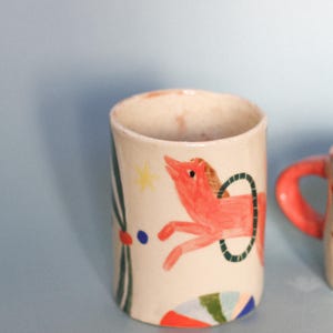 Könnte beinhalten: Eine Keramiktasse mit handgemaltem Zirkuspferdmotiv. Das Pferd ist rot mit einem grünen Reifen, einem gelben Stern und bunten Akzenten. Die Tasse hat einen cremefarbenen Boden und eine zylindrische Form.