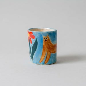 Puede incluir: Un vaso cilíndrico de cerámica con un acabado esmaltado en azul claro. El exterior presenta un gato naranja de pelo largo pintado a mano y una flor roja con hojas de color verde oscuro. El interior del vaso es de un color blanco roto neutro.