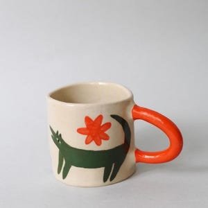Può includere: Una tazza in ceramica color crema con un animale a quattro zampe dipinto a mano in verde scuro con una lunga coda e un motivo floreale arancione brillante sopra. La tazza ha un manico spesso e arrotondato dipinto in un arancione vivace.