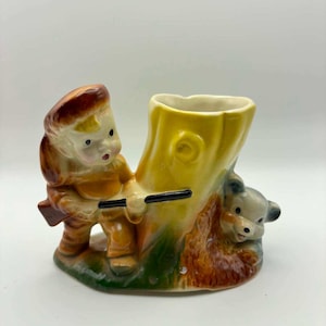 May include: A ceramic planter shaped like a tree stump featuring a small figure in a brown outfit holding a black rifle and a small bear peeking out from the base. The piece is painted in shades of yellow, brown, and green with a glossy finish.