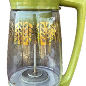May include: A vintage Sunbeam Deluxe Automatic percolator with a lime green plastic base, handle, and lid. The clear glass carafe features a gold wheat stalk pattern. A central metal stem and basket assembly are visible inside the glass container.