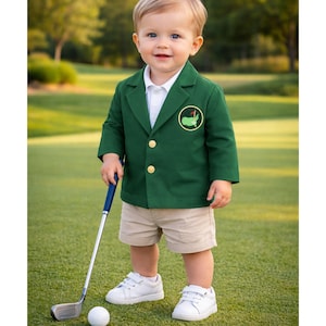 May include: A young child wearing a green blazer with gold buttons and a golf course logo, khaki shorts, and white sneakers stands on a green golf course, holding a golf club and ball. The child is smiling.
