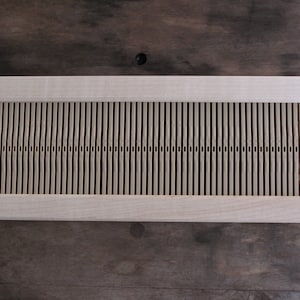 May include: A rectangular wooden weaving loom with vertical slots. The loom is made of light-colored wood and has a simple, functional design. The loom is designed for creating woven textiles.