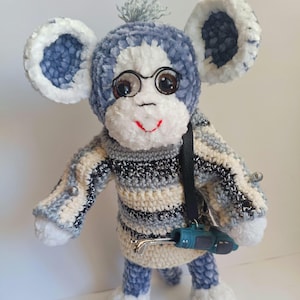 Op de afbeelding: Een handgemaakte pluche aap met een bril en een gestreepte trui. De aap heeft grote, zachte oren en houdt een miniatuur golfclub vast. De kleuren zijn voornamelijk blauw, wit en grijs, met een rode mond.