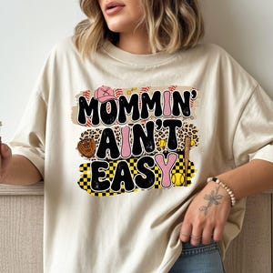 May include: Beige t-shirt featuring the text "Mommin' Ain't Easy" in a baseball-themed design. The design includes a pink baseball cap, baseballs, a leopard print section, a baseball glove, and a yellow softball.