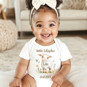 May include: A baby wearing a white onesie with a sheep graphic and the text "Gottes Schäfchen" and "Lisa". The onesie also has the date "11.12.2025" printed on it. The baby is smiling and wearing a white bow headband.