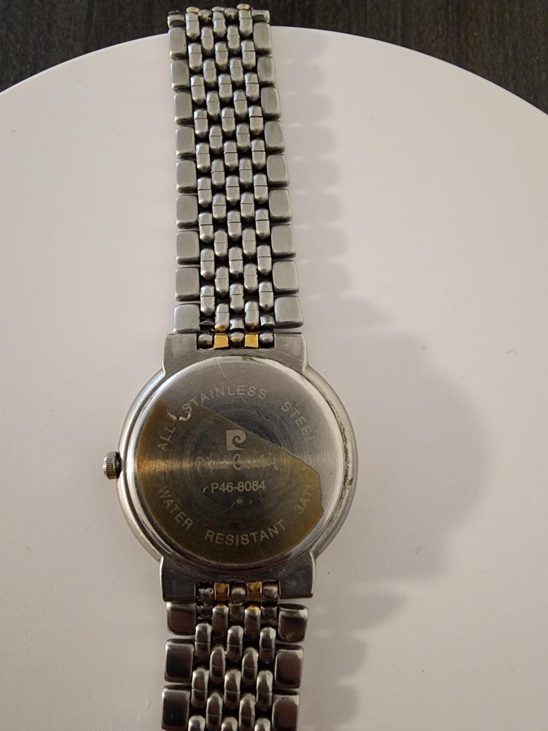 May include: A stainless steel Pierre Cardin watch with a silver band and a round watch face. The back of the watch is visible, displaying the brand name, model number P46-8084, and "Water Resistant 3ATM".