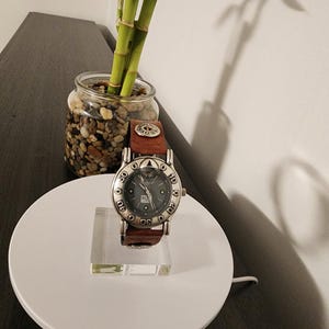 May include: A silver-toned wristwatch with a dark gray face and brown leather band. The watch is displayed on a clear acrylic stand, set on a white circular base. A small glass jar with pebbles and bamboo stalks is in the background.