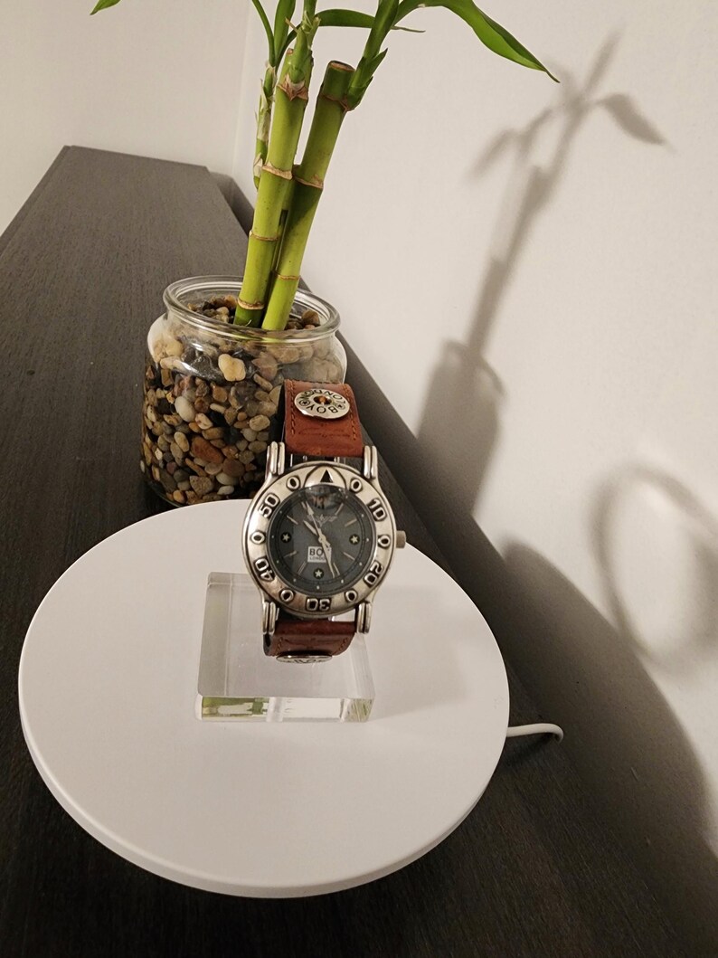 May include: A silver-toned wristwatch with a dark gray face and brown leather band. The watch is displayed on a clear acrylic stand, set on a white circular base. A small glass jar with pebbles and bamboo stalks is in the background.