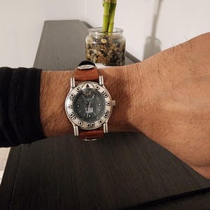 May include: A silver-toned wristwatch with a brown leather band is worn on a wrist. The watch face is dark with white numerals and a date display. The watch is on a wrist with a black sleeve.