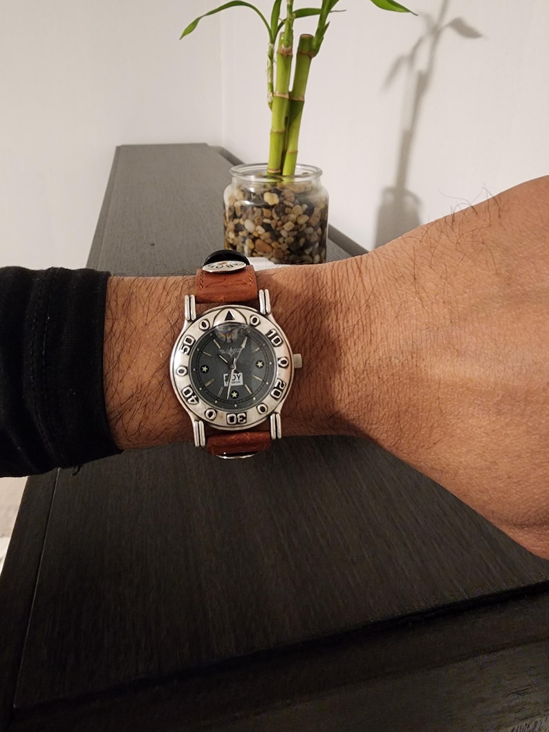 May include: A silver-toned wristwatch with a brown leather band is worn on a wrist. The watch face is dark with white numerals and a date display. The watch is on a wrist with a black sleeve.