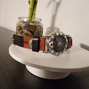 May include: A silver-toned wristwatch with a dark gray face and a brown and black leather band. The watch is displayed on a white, round platform. The band has the word "ONE" printed on it.