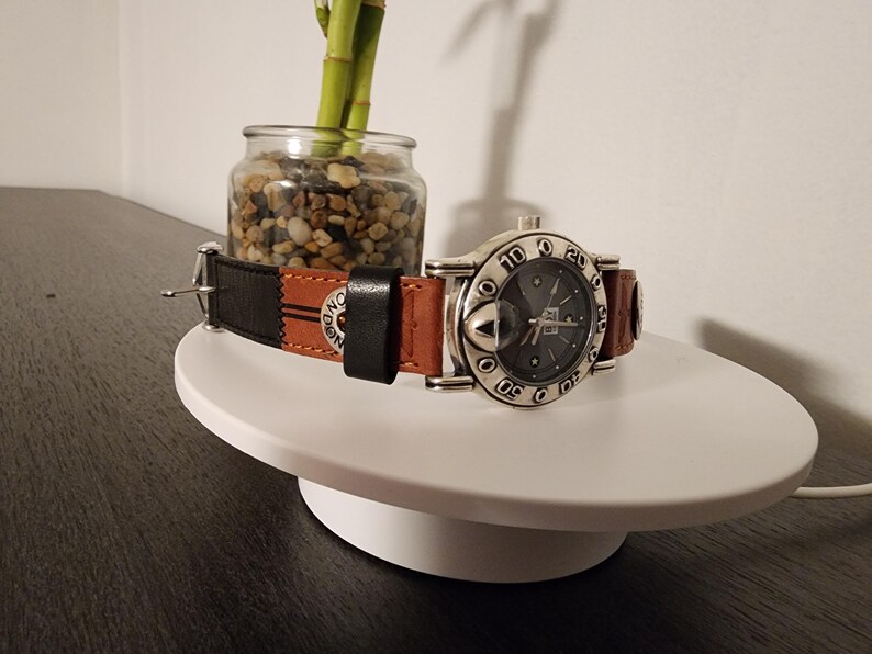 May include: A silver-toned wristwatch with a dark gray face and a brown and black leather band. The watch is displayed on a white, round platform. The band has the word "ONE" printed on it.