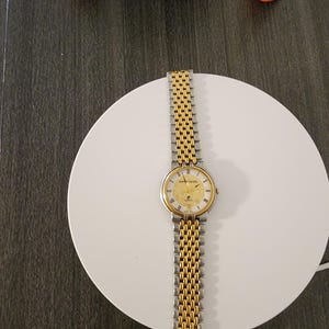 May include: A gold and silver Pierre Cardin wristwatch with a gold face and a two-tone metal band. The watch is displayed on a white circular surface. The watch face has gold hands and hour markers. The watch band is made of interlocking links.