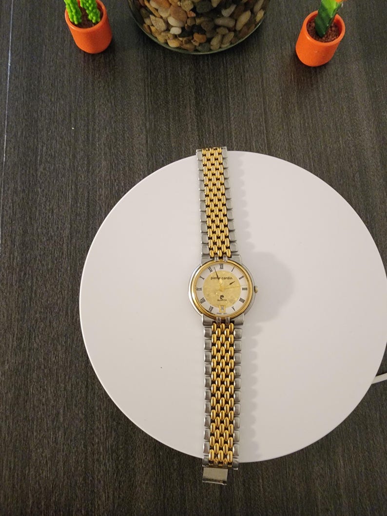 May include: A gold and silver Pierre Cardin wristwatch with a gold face and a two-tone metal band. The watch is displayed on a white circular surface. The watch face has gold hands and hour markers. The watch band is made of interlocking links.