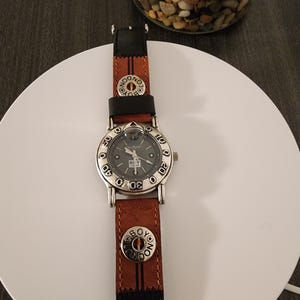 May include: A silver-toned watch with a gray face and a brown and black leather band. The watch face has the text "BOY LONDON" and the band has the same text on the button. The watch is on a white surface.