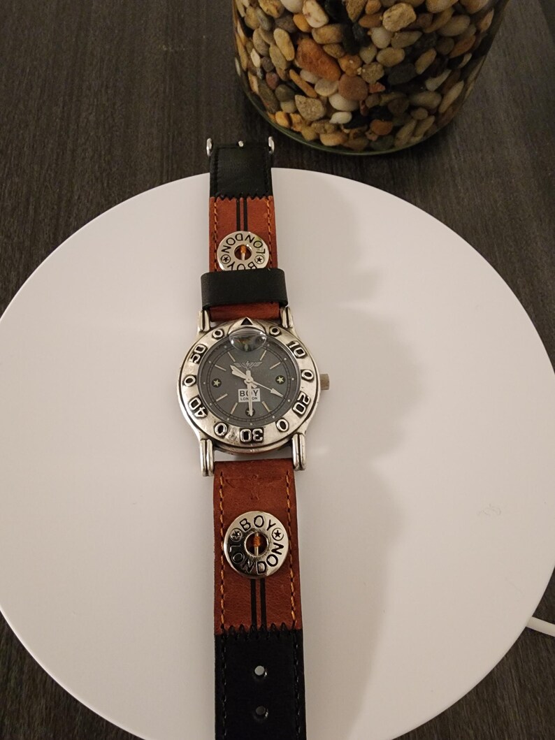 May include: A silver-toned watch with a gray face and a brown and black leather band. The watch face has the text "BOY LONDON" and the band has the same text on the button. The watch is on a white surface.