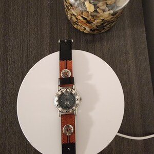 May include: A wristwatch with a brown leather band and a black leather accent. The watch face is silver with black numerals and a date display. The band features decorative metal accents. The watch is resting on a white circular surface.