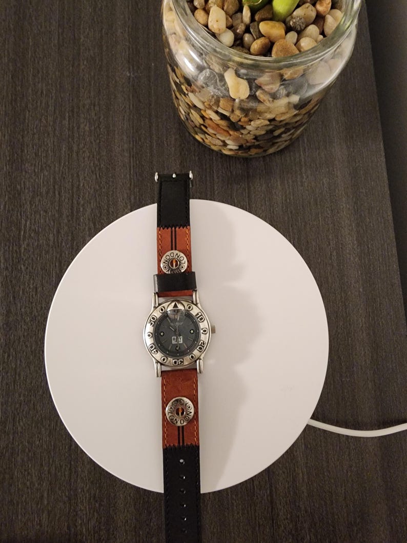 May include: A wristwatch with a brown leather band and a black leather accent. The watch face is silver with black numerals and a date display. The band features decorative metal accents. The watch is resting on a white circular surface.