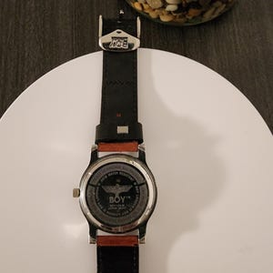 May include: Black leather watch with a silver-tone case and a red-brown accent. The watch face features the text "BOY LONDON" and "SATM WATER RESISTANT". The watch is on a white surface.