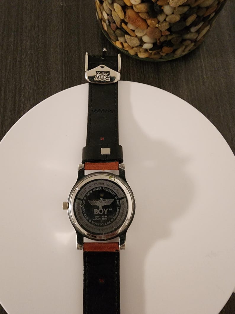 May include: Black leather watch with a silver-tone case and a red-brown accent. The watch face features the text "BOY LONDON" and "SATM WATER RESISTANT". The watch is on a white surface.