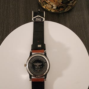 May include: A black leather watch with a silver-tone case and a brown leather accent. The watch face features the text "BOY" and a graphic. The watch is displayed on a white surface.