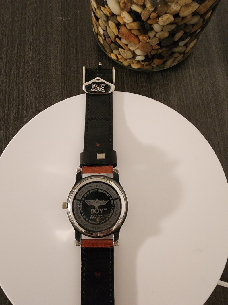 May include: A black leather watch with a silver-tone case and a brown leather accent. The watch face features the text "BOY" and a graphic. The watch is displayed on a white surface.