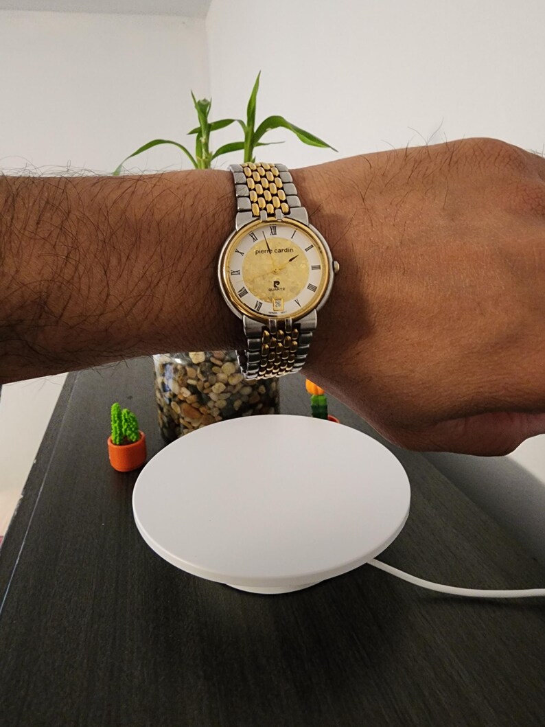 May include: A gold and silver Pierre Cardin wristwatch with a round face and a two-tone metal band. The watch is on a wrist. The watch face has gold accents and black hands. The watch is on a wrist.