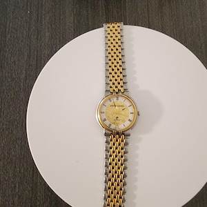 May include: A gold and silver Pierre Cardin wristwatch with a gold face and a two-tone metal band. The watch is displayed on a white plate.