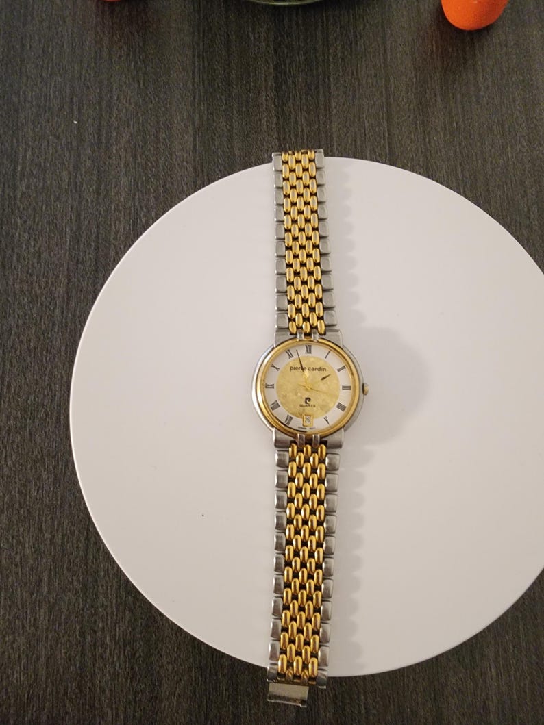 May include: A gold and silver Pierre Cardin wristwatch with a gold face and a two-tone metal band. The watch is displayed on a white plate.