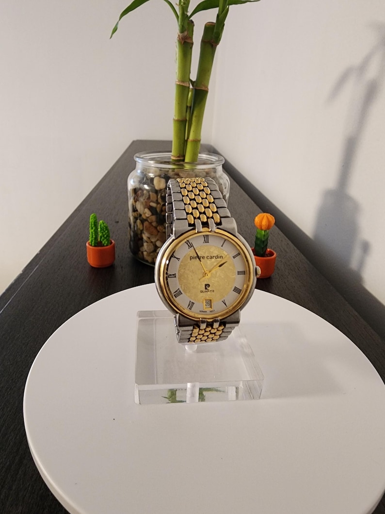 May include: A gold and silver Pierre Cardin quartz watch with a gold dial, Roman numeral hour markers, and a date window. The watch has a two-tone stainless steel band. The watch is displayed on a clear acrylic stand.