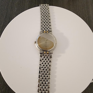 May include: A silver-toned wristwatch with a linked metal band. The watch face is round with a gold-colored bezel. The back of the watch is visible, showing the watch's inner workings. The watch is displayed on a white surface.