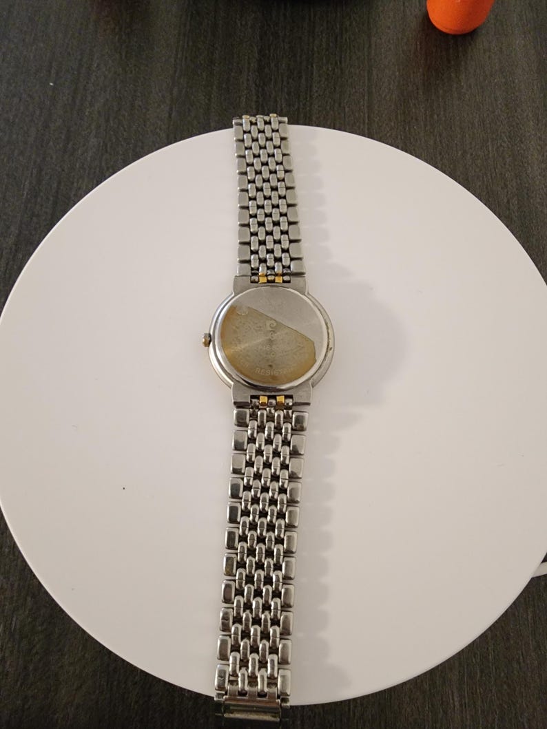 May include: A silver-toned wristwatch with a linked metal band. The watch face is round with a gold-colored bezel. The back of the watch is visible, showing the watch's inner workings. The watch is displayed on a white surface.