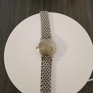 May include: A silver-toned wristwatch with a linked metal band, resting on a white circular surface. The watch face is round, and the band is composed of small, rectangular links. The watch's back is visible, showing a tarnished surface.