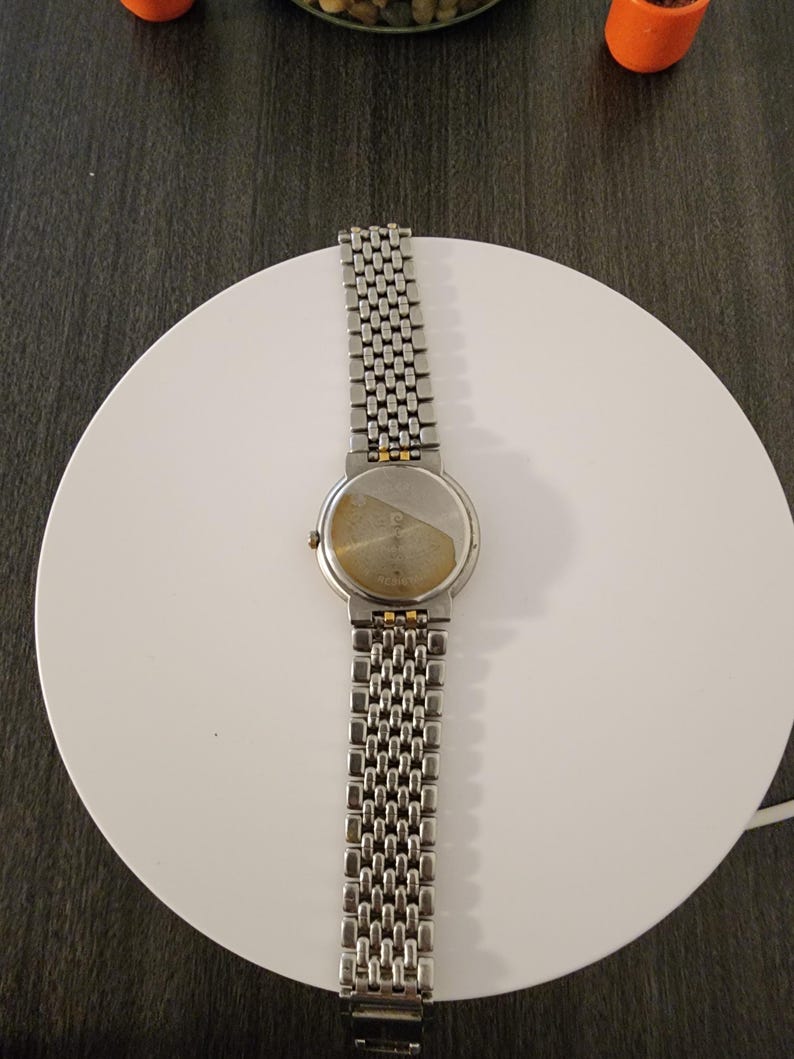 May include: A silver-toned wristwatch with a linked metal band, resting on a white circular surface. The watch face is round, and the band is composed of small, rectangular links. The watch's back is visible, showing a tarnished surface.