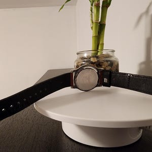 May include: A black leather watch band with a silver buckle is displayed. The watch face is silver with a black leather band. The watch is on a white surface with a small glass jar of plants in the background.