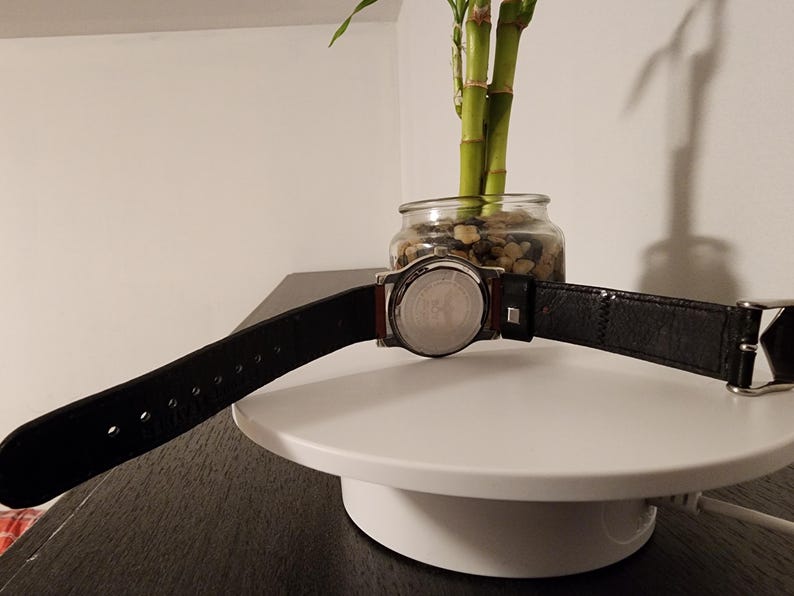 May include: A black leather watch band with a silver buckle is displayed. The watch face is silver with a black leather band. The watch is on a white surface with a small glass jar of plants in the background.