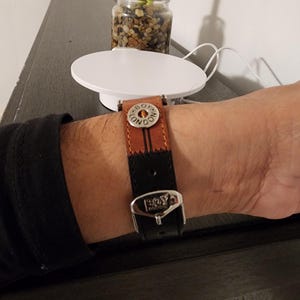 May include: A brown and black leather watch band with a silver buckle. The band has the words "BOY LONDON" on the buckle and a decorative button. The watch band is on a wrist.
