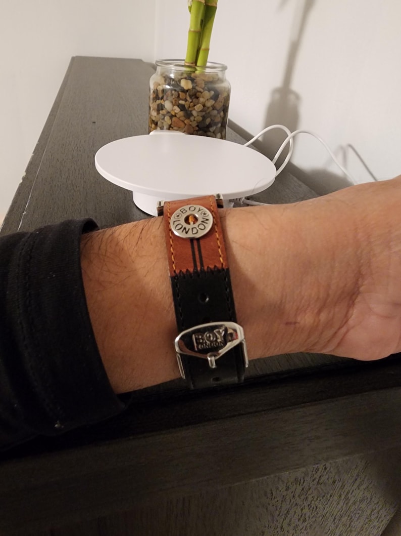 May include: A brown and black leather watch band with a silver buckle. The band has the words "BOY LONDON" on the buckle and a decorative button. The watch band is on a wrist.
