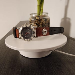 May include: A silver-toned wristwatch with a dark gray face and a brown and black leather band. The watch is displayed on a white, circular platform. The watch face has numbers and markers.