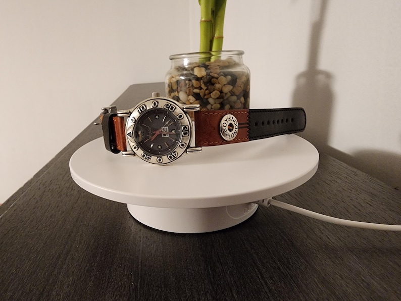 May include: A silver-toned wristwatch with a dark gray face and a brown and black leather band. The watch is displayed on a white, circular platform. The watch face has numbers and markers.