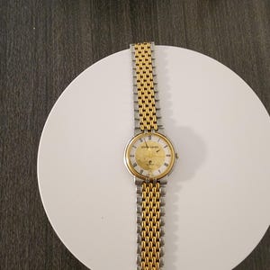 May include: A gold and silver-toned Pierre Cardin wristwatch. The watch has a gold face with black markers and hands. The band is a combination of gold and silver links. The watch is displayed on a white surface.